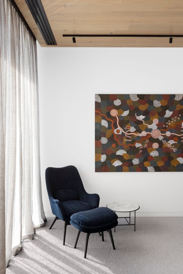A refined sitting area with a deep navy upholstered lounge chair and matching ottoman beside a small round marble-topped side table. Full-height sheer curtains cover a large window, while a colourful abstract artwork hangs on a white wall. The room features pale timber ceiling lining, black recessed track lighting, and a light grey carpet, creating a calm contemporary interior with mid-century-inspired furniture.