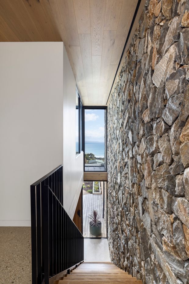Interior view down a narrow stairway beside a dramatic exposed rough-stone feature wall. The stair has warm timber treads and a slim black metal balustrade, with pale timber ceiling panelling, white walls, and dark-framed glazing at the end revealing a coastal ocean outlook and a lower deck. The space is bright, contemporary, and materially focused.