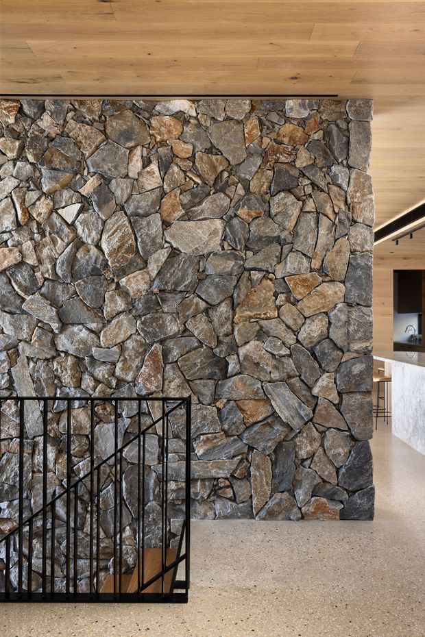 Interior architectural detail featuring a tall, irregular natural-stone feature wall beside a black metal stair balustrade. Warm timber ceiling and flooring frame the stonework, while a polished light terrazzo floor extends toward an open kitchen or living area in the background.