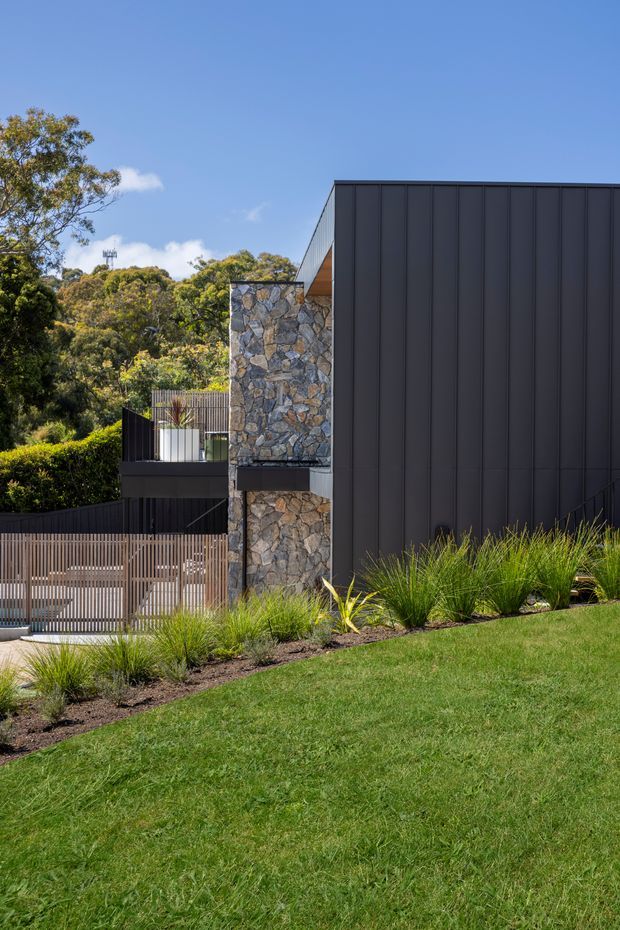 Exterior view of a contemporary residential building on a landscaped, sloping site. The facade combines dark vertical metal cladding with prominent irregular natural stone walls, while a slatted metal gate and balcony fencing sit among layered garden planting and lawn. Bright clear daylight and surrounding trees frame the building.
