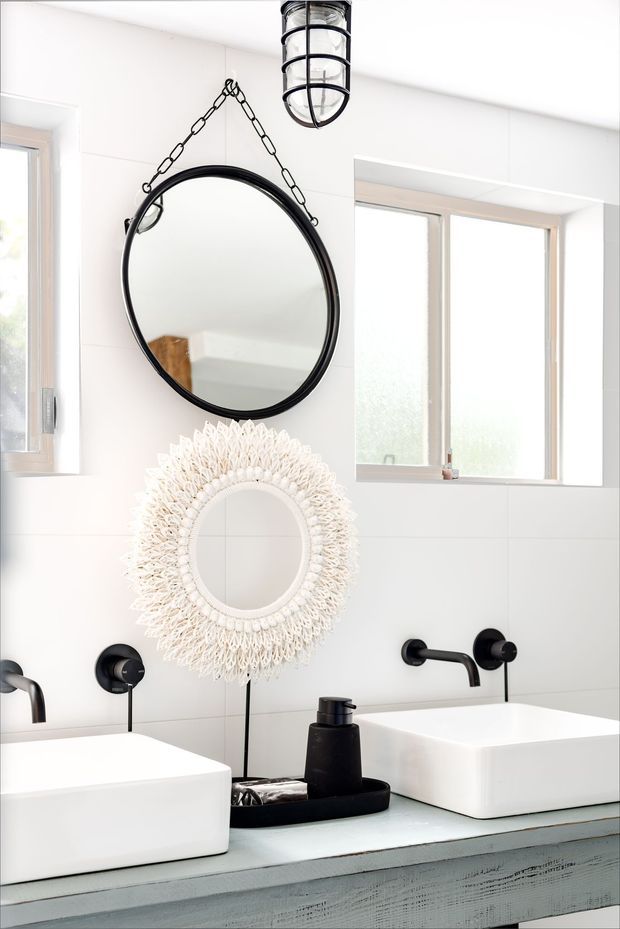 Bright contemporary bathroom vanity area with twin white vessel basins on a weathered pale blue-grey timber vanity, white tiled walls, black wall-mounted tapware, large round black-framed mirror, decorative woven circular mirror, and black caged pendant light. Natural light enters through frosted windows, creating a clean high-contrast interior.