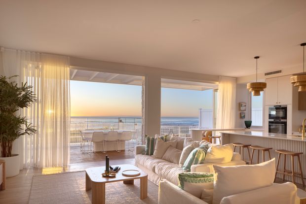 Bright open-plan coastal living and kitchen area with a large cream sectional sofa, timber coffee table, white kitchen island with bar seating, and full-height glazed openings leading to a balcony overlooking the ocean. Sheer curtains, warm neutral finishes, timber accents, and pendant lights create a relaxed, airy atmosphere.