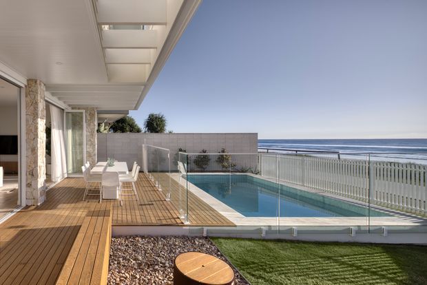A bright coastal terrace overlooking the ocean, with a timber deck, glass balustrades, and a rectangular swimming pool enclosed by pale tiled walls and white metal fencing. The covered outdoor area includes a dining setting and sliding glass doors opening to the home. The scene has a clean, contemporary resort-like character with strong horizontal lines and expansive ocean views.