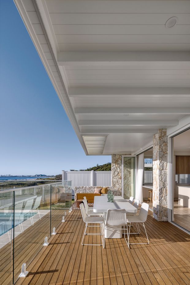A bright coastal outdoor dining terrace with a wide white-lined soffit and exposed beams overhead, natural stone-clad columns, and large sliding glass doors opening into the interior. The terrace has warm timber decking, a glass balustrade, a white dining table with modern outdoor chairs, and built-in seating. A coastal water view is visible beyond the property.