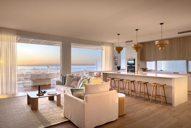 Bright open-plan coastal living space with a contemporary kitchen and island breakfast bar opening onto a furnished balcony or terrace. The room features large glazed openings with sheer curtains, a soft neutral lounge arrangement, light timber flooring, warm wood cabinetry, and three woven pendant lights. An ocean horizon is visible beyond the terrace.