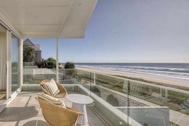A bright beachfront balcony or terrace viewed from beside a coastal home, with frameless glass balustrades, pale decking, woven outdoor lounge chairs, and a small round table. The balcony overlooks a sandy beach and ocean, while a neighbouring coastal house is visible to the side. The architecture is clean-lined and contemporary with a broad flat soffit and large glazed openings.