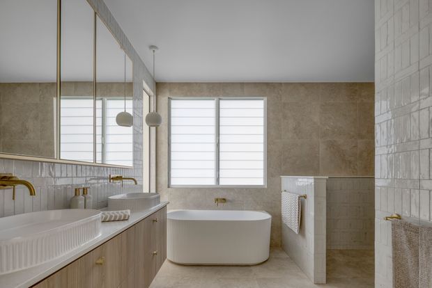 A bright, refined contemporary bathroom with a freestanding white bathtub beneath large horizontal windows. The room includes a double vanity with timber cabinetry, wall-to-wall mirrors, brass tapware, white glazed wall tiles, warm stone-look wall and floor finishes, and a partially enclosed shower area. Soft natural light creates a calm, spa-like atmosphere.