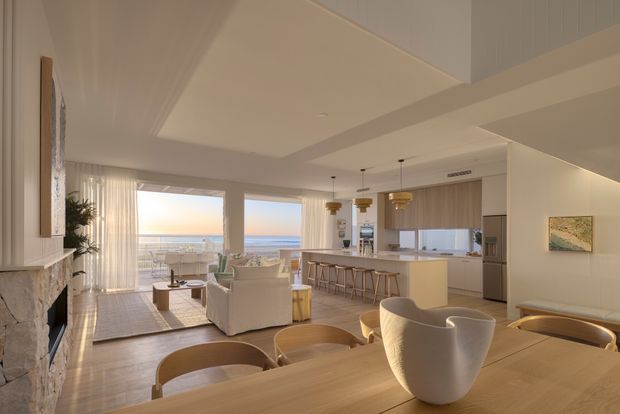 Bright open-plan coastal apartment interior with a living room, dining area, and kitchen arranged around expansive ocean-facing glazing. The space features a light timber dining table and flooring, pale upholstered seating, a stone fireplace, integrated kitchen cabinetry, pendant lights, and a balcony or terrace visible beyond the living area. A large ceiling bulkhead and open upper volume add architectural interest.