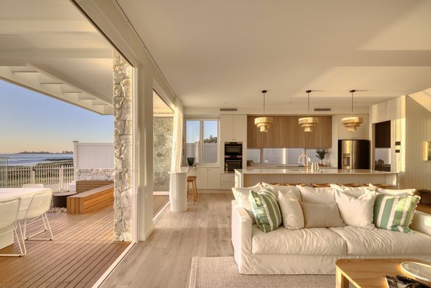 An airy open-plan coastal living space with a white slipcovered sofa in the foreground, a light-toned kitchen with timber cabinetry and a stone island behind it, and a dining area opening onto a timber deck. Large glazed openings connect the interior to a waterfront-facing outdoor terrace, while white stone-clad columns and warm layered pendant lighting add texture and refinement.