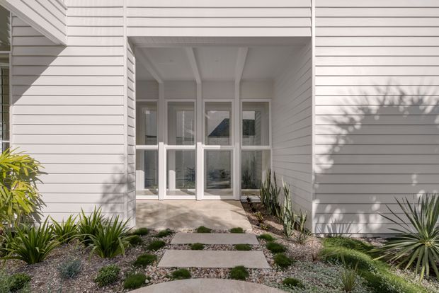 Front entry of a contemporary single-storey home with pale horizontal weatherboard cladding, a recessed porch framed by white posts and beams, and a glazed double-door entrance. A low-maintenance landscaped path of large concrete stepping slabs, gravel, succulents, and grasses leads to the doorway. Strong sunlight creates crisp shadows across the facade.