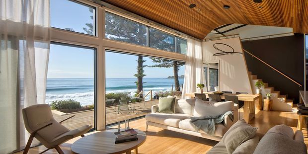 Bright contemporary living room with expansive floor-to-ceiling glazing overlooking the ocean. The room features a warm timber slatted ceiling, polished timber flooring, a large cream sectional sofa, sculptural lounge chair, round coffee table, and a dramatic black staircase with light timber steps and minimalist balustrade. Sheer curtains soften the strong coastal light, while a deck and beach are visible outside.