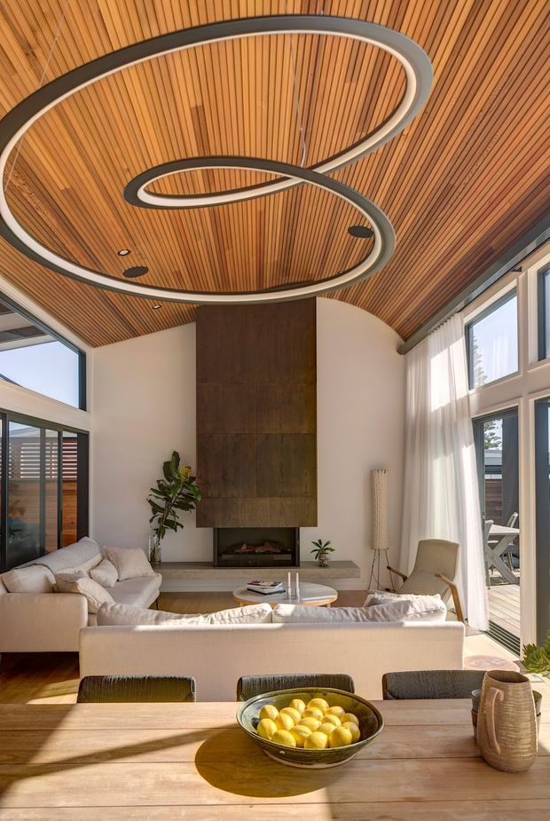 A bright, refined open-plan living and dining area with a dramatic curved timber-lined ceiling and sculptural looping pendant light. The room features a large cream sectional sofa, a tall dark timber fireplace surround, expansive glazing with sheer curtains, and a timber dining table in the foreground. A deck is visible through the doors.