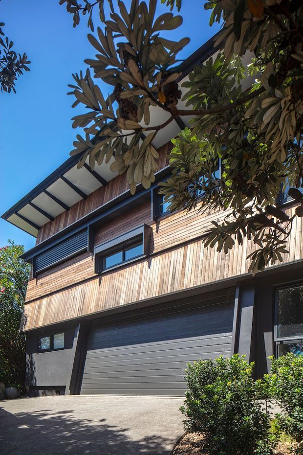 Street-level exterior view of a modern multi-storey residence, dominated by a large grey garage door, dark structural framing, and horizontal timber cladding. The steeply pitched roof has broad eaves, while mature leafy vegetation partially frames the facade. Bright daylight and a clear blue sky are visible.