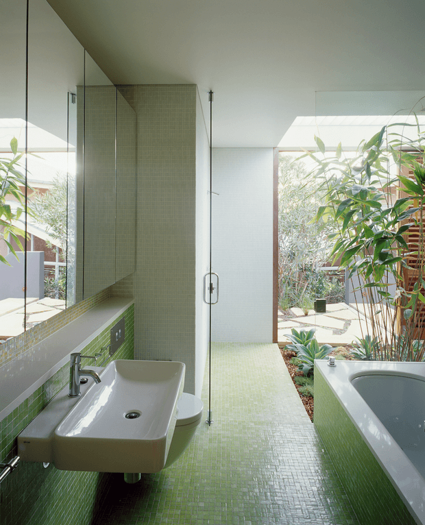 Bright contemporary bathroom with a wall-mounted rectangular basin, chrome tapware, glass shower enclosure, freestanding bathtub, and extensive small-format green mosaic tile flooring and wall detailing. A large mirror reflects the basin area, while full-height glazing opens views toward a planted courtyard.