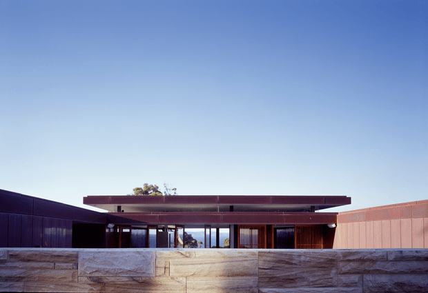 Low-slung modern single-storey building viewed from the exterior, with a broad flat roof, deep horizontal overhangs, extensive floor-to-ceiling glazing, dark vertical screening, and a textured stone-clad boundary wall. The composition is minimalist and strongly horizontal beneath a clear blue sky.