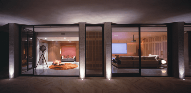 A dark, low-profile modern residential interior viewed through a full-height glazed facade at night. The composition shows two connected living areas behind black-framed glass: a warmly lit lounge with a large sectional sofa, television, timber wall panelling and a suspended ceiling fan, and an adjacent sitting area with a sculptural floor lamp, low seating and a round textured rug. Strong vertical structural piers, concealed uplighting and a dark ceiling create a dramatic, gallery-like atmosphere.