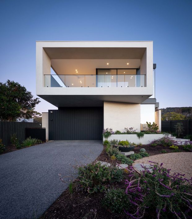 Contemporary two-storey residential house photographed from the front at dusk. The building has a crisp white rendered rectangular upper volume with a deep recessed balcony, clear glass balustrade, and extensive dark-framed glazing. A projecting upper floor shelters a dark vertical-panel garage, while the lower level combines pale masonry/rendered walls with landscaped garden beds, gravel, and a paved driveway. The composition highlights the modern facade, balcony, garage, and low-maintenance front landscaping against a blue evening sky.