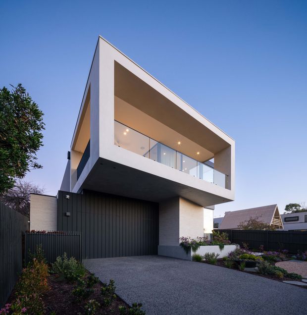 Contemporary two-storey residence viewed from the street, featuring a bold cantilevered upper level with a deep recessed balcony, frameless glass balustrade, white rendered walls, dark vertical garage and boundary cladding, and a landscaped gravel driveway and garden.