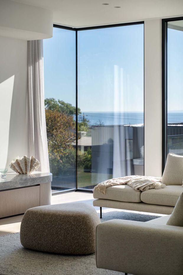 A bright, refined living room with floor-to-ceiling corner glazing overlooking the ocean. The space features a cream upholstered sectional sofa, rounded ottoman, textured light rug, pale stone-topped timber console, sheer curtains, and minimal white walls and ceiling. Strong natural daylight and black-framed windows create a calm contemporary coastal atmosphere.