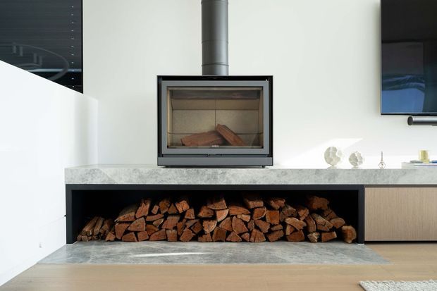 Modern living room feature wall with a built-in wood-burning fireplace set on a pale stone hearth and benchtop, with neatly stacked firewood stored in an open niche below. Light timber cabinetry, pale walls, and a wall-mounted television create a clean contemporary interior.