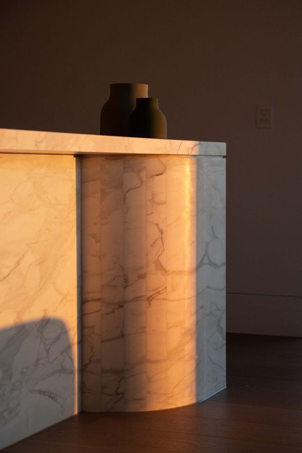 Close-up interior detail of a curved, freestanding marble-clad built-in feature or cabinet with pronounced veining, illuminated by warm sunlight. Dark timber flooring and a deep taupe wall create a moody backdrop, while two matte black decorative vessels sit on top. The image emphasizes sculptural joinery, stone texture, and dramatic light and shadow.