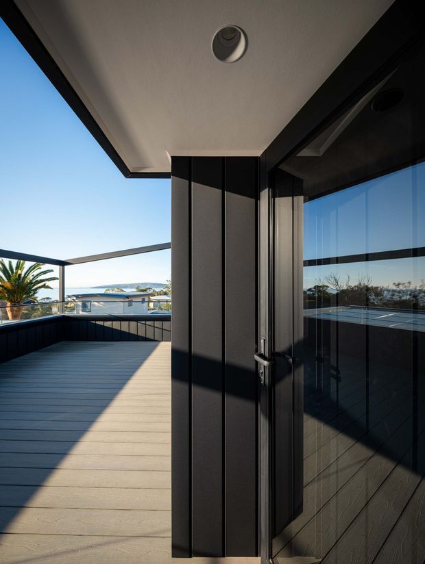 A modern covered outdoor terrace or balcony with pale composite decking, dark vertical cladding and black-framed glass doors. The terrace has glass balustrades and overlooks a coastal residential setting with water visible in the distance. Strong sunlight, clean lines and reflective glazing create a contemporary architectural feel.