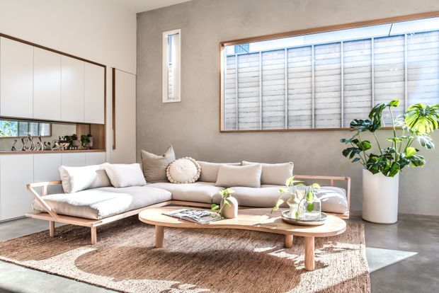 Bright, refined living room with a low-profile pale sectional sofa, sculptural timber coffee table, textured neutral rug, indoor plants, and extensive built-in cabinetry. A large horizontal window with external screening and a narrow vertical window bring in abundant natural light. The space has a warm neutral palette and clean-lined mid-century-inspired furnishings.