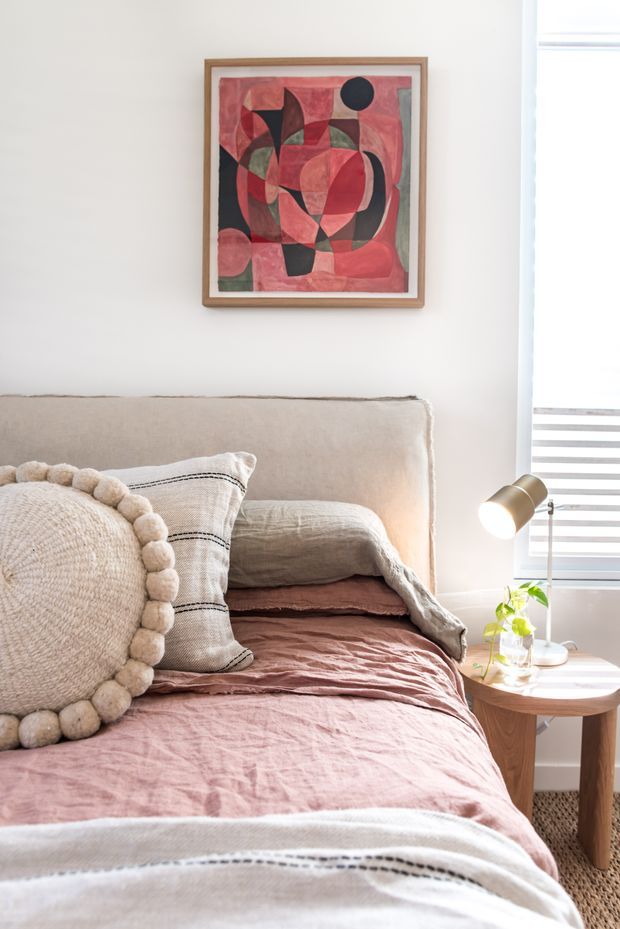 A softly lit bedroom vignette focused on a neatly made bed with a textured upholstered headboard, layered blush-pink and beige bedding, and decorative neutral cushions. A small round timber side table holds a brass-toned lamp and a small plant, while an abstract geometric artwork hangs above the bed. White walls, bright window blinds, and a textured carpet create a warm, relaxed atmosphere.