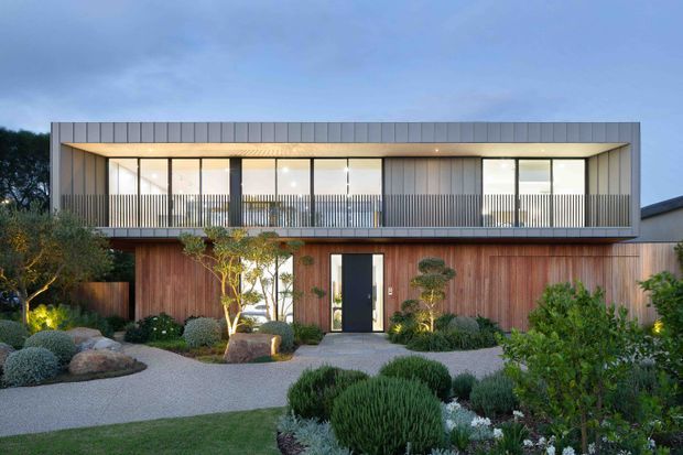 A contemporary two-storey residence with a long rectangular form, vertical timber-clad lower level, and a recessed upper level framed in grey metal cladding. Full-height glazing opens onto a balcony with a slender vertical balustrade. The entrance is set within landscaped native-style gardens featuring gravel paths, sculptural rocks, clipped shrubs, and low planting.