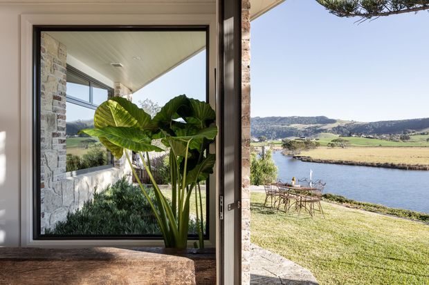 View from a contemporary home through a large glazed opening toward a landscaped waterfront setting. The foreground includes a stone-clad wall, pale timber-lined ceiling, dark window and door frames, and a substantial natural stone ledge or bench, with large leafy plants framing the view. Beyond is a broad body of water, grassy shoreline, rural fields, and rolling hills.