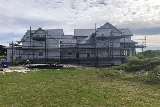 Exterior view of a large multi-gabled house under construction, with light weatherboard-style cladding, pitched metal roofs, extensive scaffolding, and an open grassy site in the foreground.