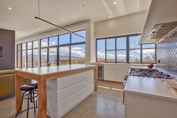 Bright contemporary kitchen with a large timber-framed island and white drawers, integrated cooktop and cabinetry, patterned tile splashback, polished concrete-style flooring, and extensive black-framed windows overlooking a mountain landscape. A linear pendant light hangs above the island, with bar stools providing seating.
