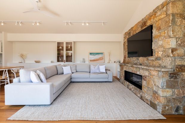 Bright, open-plan living room with a large light-grey sectional sofa, textured neutral rug, built-in shelving and cabinetry, and a substantial exposed natural-stone fireplace wall incorporating a recessed television and fireplace. A dining or kitchen area is partially visible at the left, with contemporary track lighting and a ceiling fan.