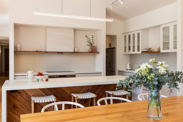 Bright contemporary kitchen and dining area with a large white island, integrated cooktop, pale tiled splashback, white cabinetry with glass-front doors, timber shelving, and a warm timber dining table. White spindle-back dining chairs and woven stools add a relaxed, refined character. The room has a high sloping ceiling with track lighting and a long linear pendant over the island.