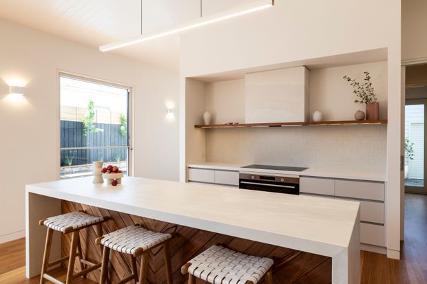 Bright contemporary kitchen with a large white waterfall-edge island, integrated cabinetry, induction cooktop, built-in oven, pale textured backsplash, and a timber open shelf with restrained decor. Three woven-seat timber stools line the island, while large glazing and warm timber flooring add natural warmth.