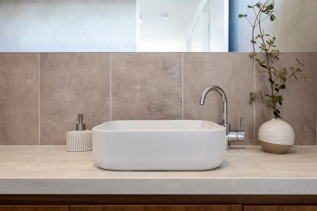 Close-up of a contemporary bathroom vanity featuring a white rectangular vessel basin, a curved chrome mixer tap, beige-grey large-format tiled splashback, pale stone-look countertop, and dark timber cabinetry below. A mirror and small decorative plant are visible in the background.