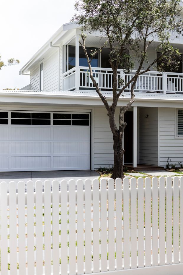 A bright two-storey residential house facade viewed from the street, featuring white horizontal weatherboard cladding, a white garage door, covered upper-level balcony with white vertical balustrades, and a white picket fence in the foreground. A mature tree partially obscures the facade, while a concrete driveway and front entry are visible.