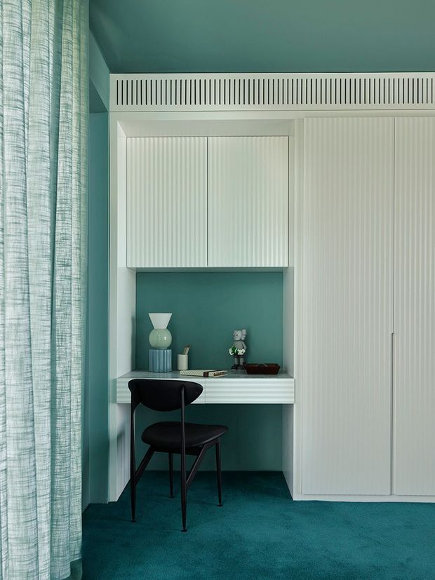 A compact built-in home office nook set within a teal room. White fluted cabinetry and a matching built-in desk surround an open shelf displaying small decorative objects, with a black sculptural chair positioned at the desk. Teal carpeting, teal walls and ceiling, and a sheer textured curtain create a cohesive monochromatic interior with strong vertical ribbed detailing.