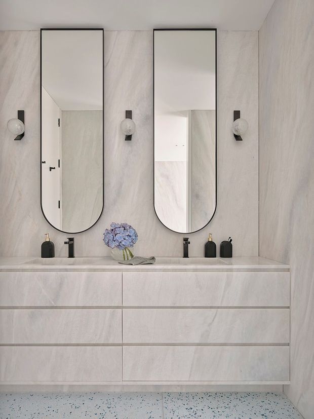 A refined contemporary bathroom vanity with a wide pale stone-look double vanity, integrated or closely matched benchtop, two black-framed rounded-rectangle mirrors, minimalist black wall sconces, twin black tapware, and dark accessories. The walls and cabinetry share a soft veined light finish, while the floor is terrazzo with small dark and blue-grey chips.