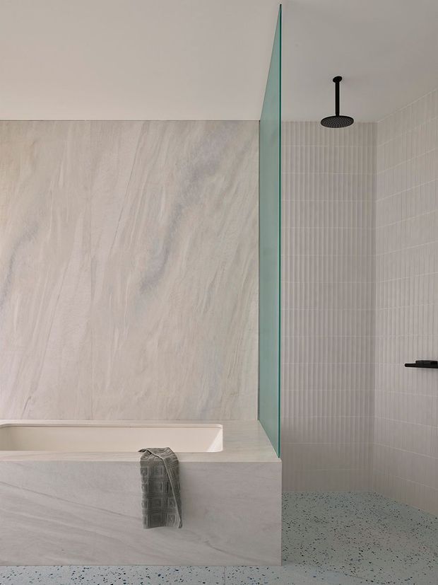 Minimalist contemporary bathroom featuring a freestanding-style built-in bathtub clad in pale veined stone-look material, a walk-in shower separated by a clear frameless glass panel, fine vertical wall tiles, black shower fittings, and a terrazzo-like speckled floor. The palette is soft, neutral, and refined.