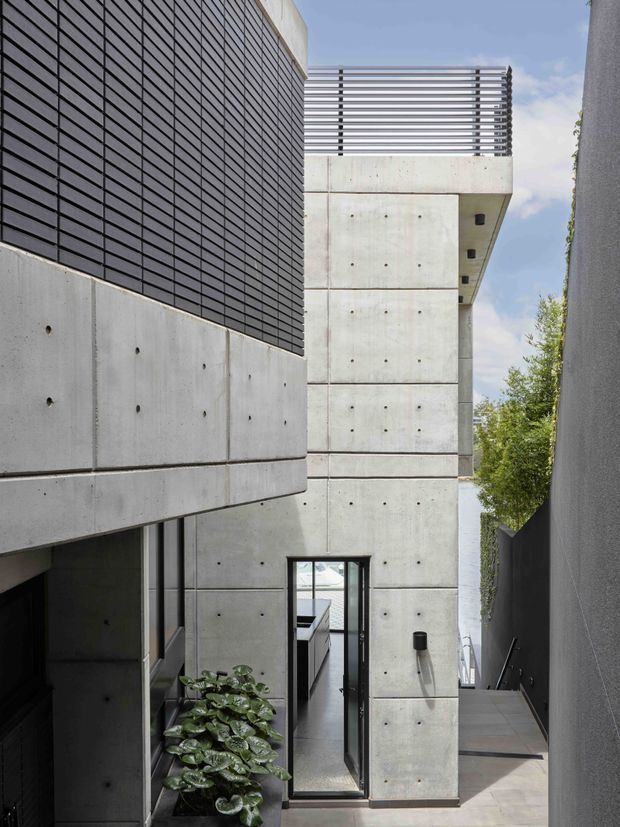 Narrow modern architectural exterior courtyard or side passage framed by large exposed concrete walls and panels, with a dark horizontal slatted screen on the left, a metal-railed upper balcony, black-framed glazing and a tiled walkway. Strong geometric composition, raw concrete detailing, and planted greenery visible along the passage.