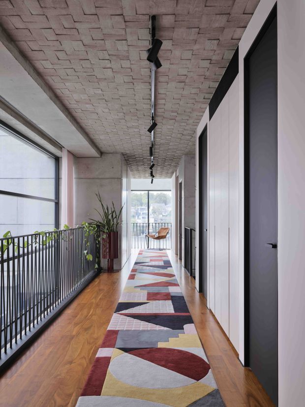 A long, bright interior corridor or landing with warm timber flooring, a bold geometric runner rug, white built-in or paneled doors on one side, and a black metal balustrade overlooking a lower level on the other. The ceiling features exposed pale brickwork in a repeating pattern with a linear track of black spotlights. Large windows provide abundant natural light, and potted plants add greenery near the far end.