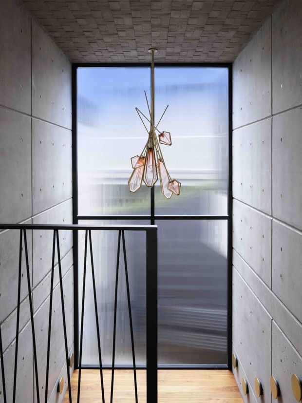A narrow modern stair landing with raw concrete walls and a patterned concrete ceiling. Black metal balustrading frames a timber stair or landing floor, while a large translucent floor-to-ceiling window provides diffuse daylight. A sculptural geometric pendant hangs in front of the glazing.