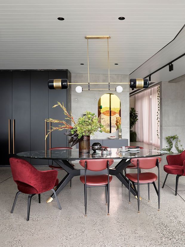 A sophisticated contemporary dining area with a long glass-top dining table surrounded by sculptural red upholstered chairs. The room features a dark built-in cabinet wall, polished terrazzo flooring, a grey concrete-look wall, a white linear-panel ceiling, large glazing with sheer pink curtains, and a striking geometric pendant light. Decorative greenery, floral arrangements, and a circular artwork add colour and visual interest.