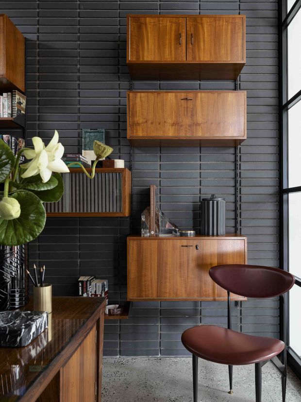 A refined mid-century-inspired interior vignette featuring walnut-toned floating cabinetry and shelving mounted against a dark charcoal brick wall. The space includes a timber sideboard, vintage-style leather chairs, books, decorative objects, indoor plants, and a large window providing strong natural light. The polished timber joinery contrasts with the dark masonry backdrop and terrazzo-like concrete floor.