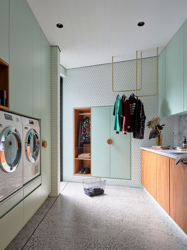 A bright, contemporary laundry room with stacked front-loading washer and dryer integrated into pale green cabinetry on the left. The room features a terrazzo floor, mint-green wall panels and cabinetry, a white hexagonal-tile backsplash, a timber-faced laundry bench with a pale stone benchtop, open wardrobe storage, hanging rails, and a utility sink area on the right.
