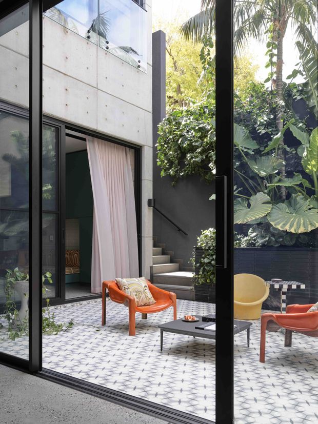View through large black-framed glazing into a landscaped, enclosed courtyard. The courtyard has patterned tiled paving, contemporary outdoor lounge chairs in orange and yellow, a low dark coffee table, dense tropical planting, and concrete architectural walls with an external stair. A curtained opening leads into the adjoining building.