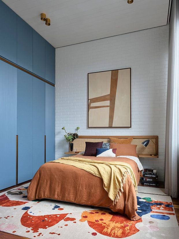 A warmly styled bedroom with a high white painted brick feature wall, full-height blue built-in wardrobes with slim timber handles, and a timber-framed upholstered headboard with integrated bedside shelving. The bed is dressed in rust, terracotta, mustard and patterned textiles, positioned over a colourful abstract area rug. A large abstract artwork, small wall-mounted reading lights, and sheer curtains complete the contemporary interior.