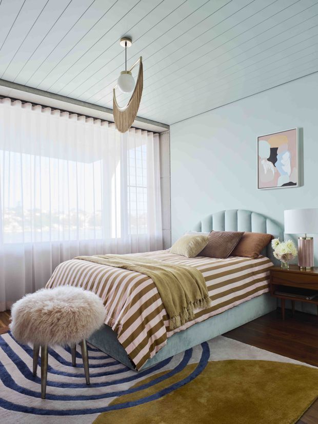 Bright, softly styled bedroom with a low upholstered bed, vertically channelled pale blue-green headboard, striped brown-and-cream bedding, and layered olive and neutral throws. A large sheer-curtained window provides abundant daylight, while a sculptural pendant hangs from a white-lined ceiling. The room includes a dark timber bedside table, table lamp, abstract wall art, a fluffy stool, and layered curved-pattern rugs over dark timber flooring.