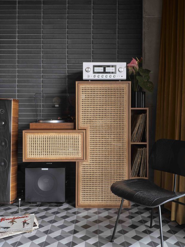 A moody, richly layered music listening corner with vintage-inspired audio equipment, including a turntable, silver stereo receiver, large cane-front speakers, and a black subwoofer. The setup sits against a dark charcoal brick-tile wall beside open vinyl storage shelving. A black leather lounge chair, heavy brown curtains, patterned geometric floor tiles, and a leafy plant complete the retro, tactile interior.